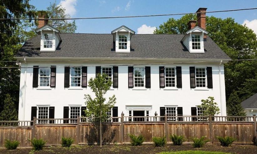 White two-story house with black shutters, dormer windows, and a wooden fence in front.