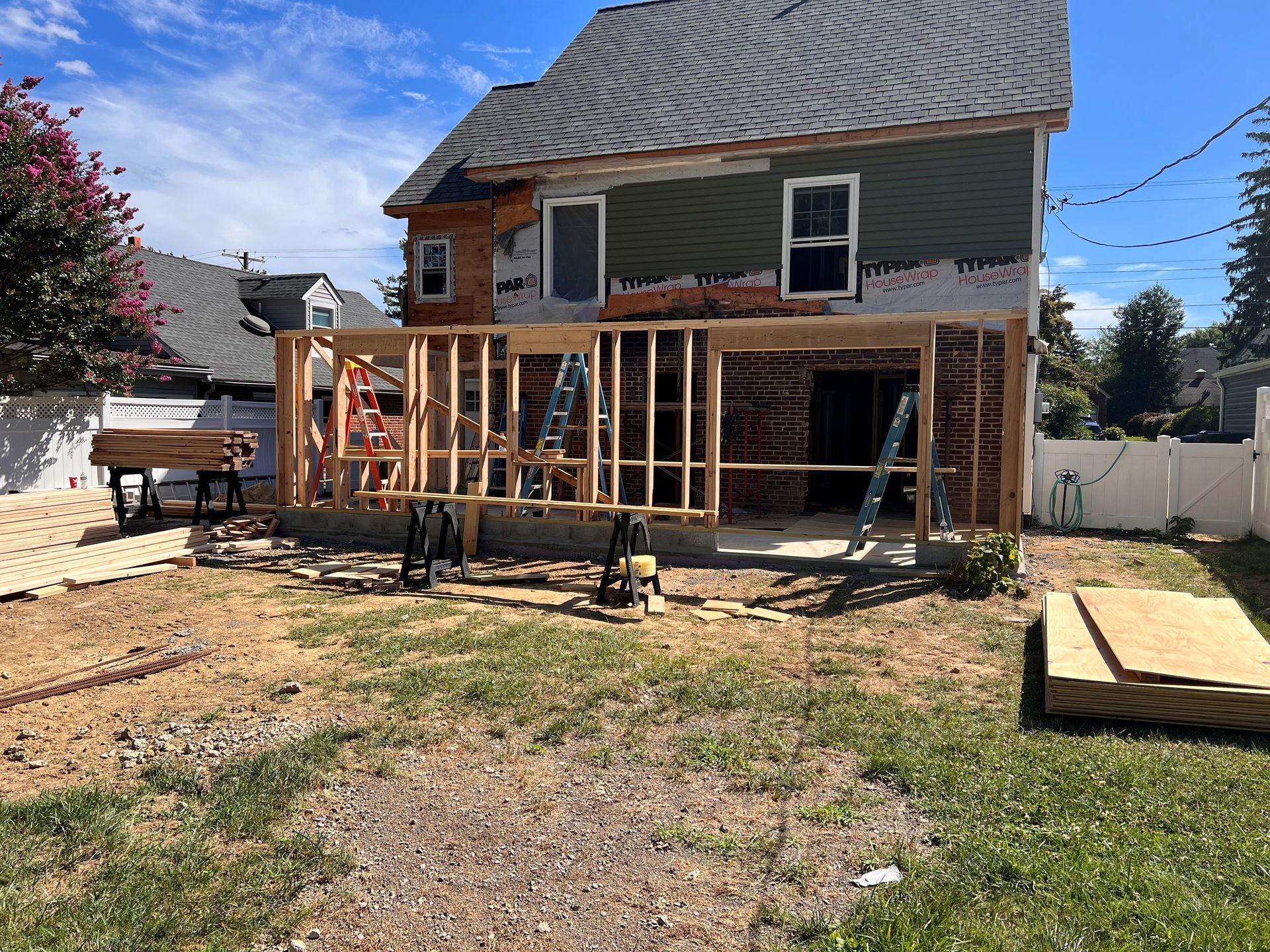 Construction of a wooden frame extension to a two-story house. Dry grass yard, blue sky.