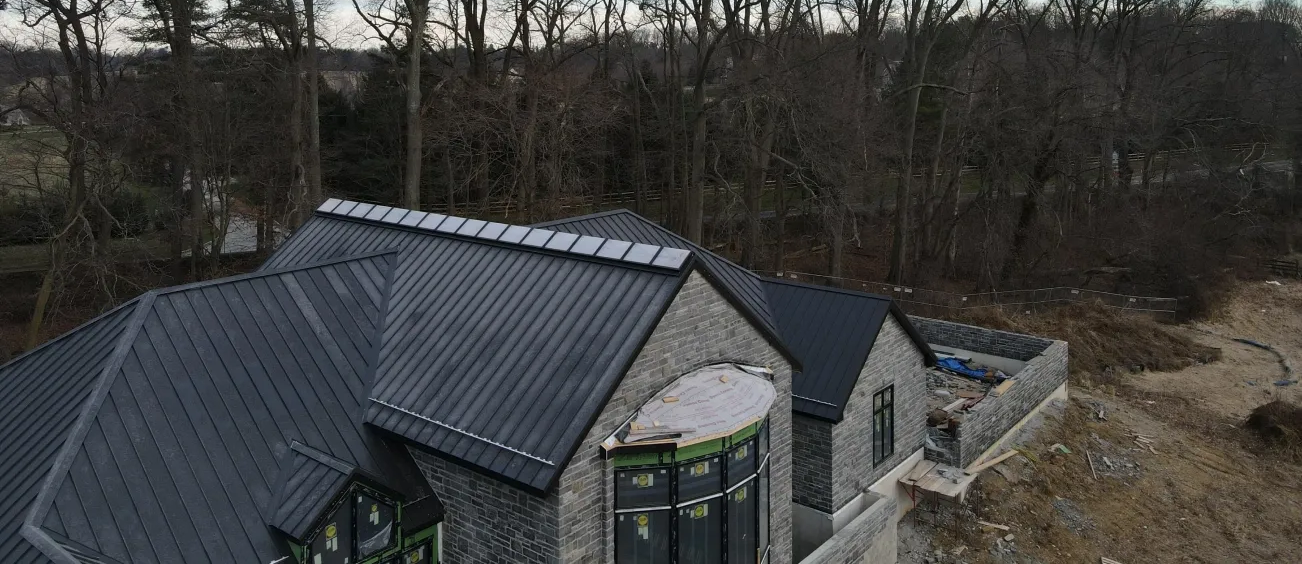 Aerial view of a house with a dark metal roof, stone walls, and surrounding trees.