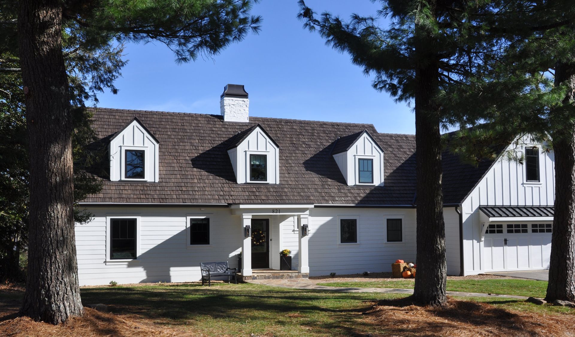 White house with brown shingle roof, three dormer windows, black framed windows, and a garage, framed by trees.