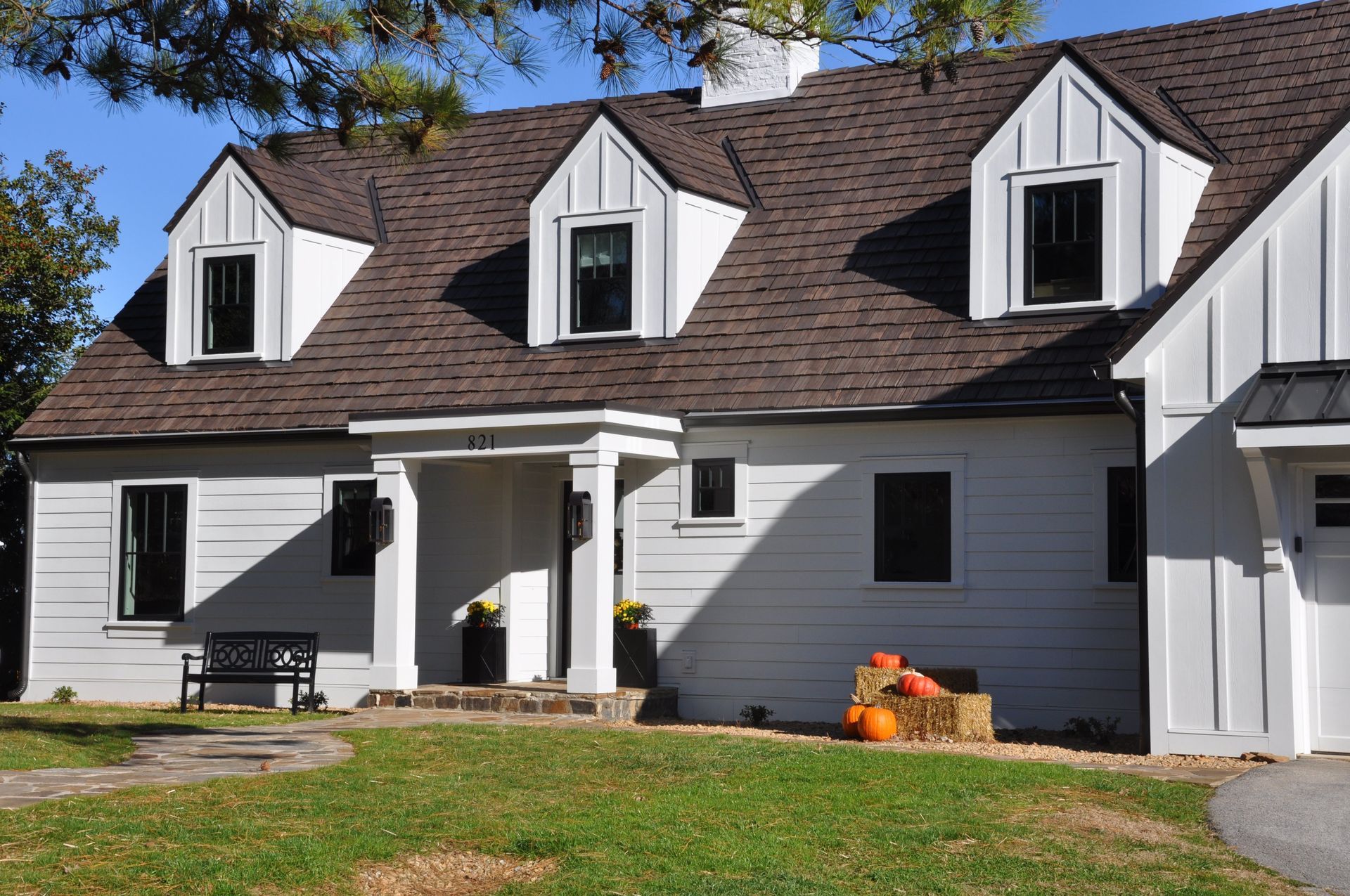 White house with three dormers, dark roof, black windows, pumpkins on a bale of hay in yard.