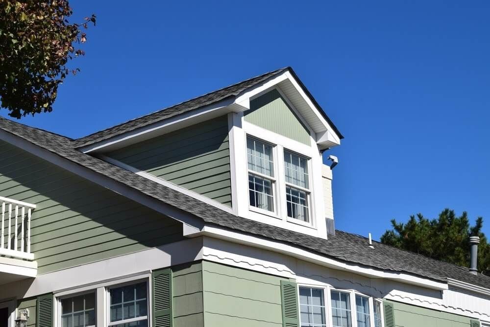 Green house with white trim, dormer with windows, and a blue sky.