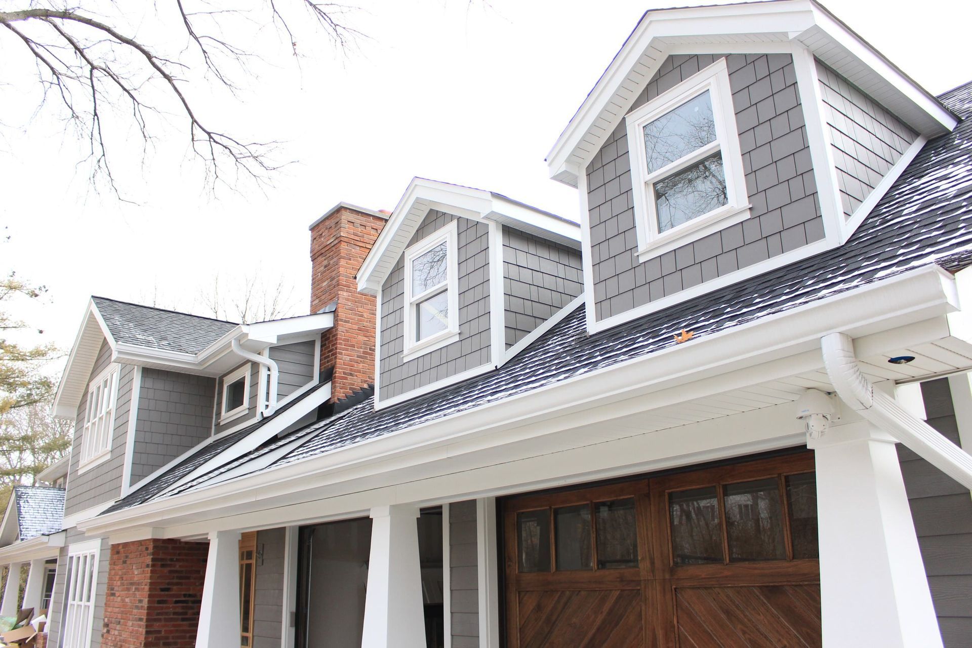 Gray house with white trim, three dormers, black roof, and wooden garage doors.