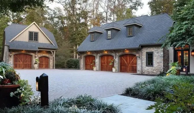 Stone garage with wooden doors and dormers, next to a detached storage building, on a brick driveway.