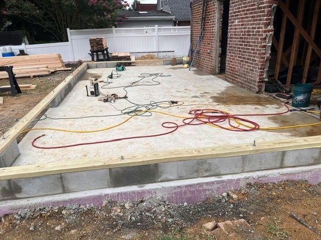 Construction site: Concrete slab with lumber frame, cinder blocks, tools, and exposed brick wall.