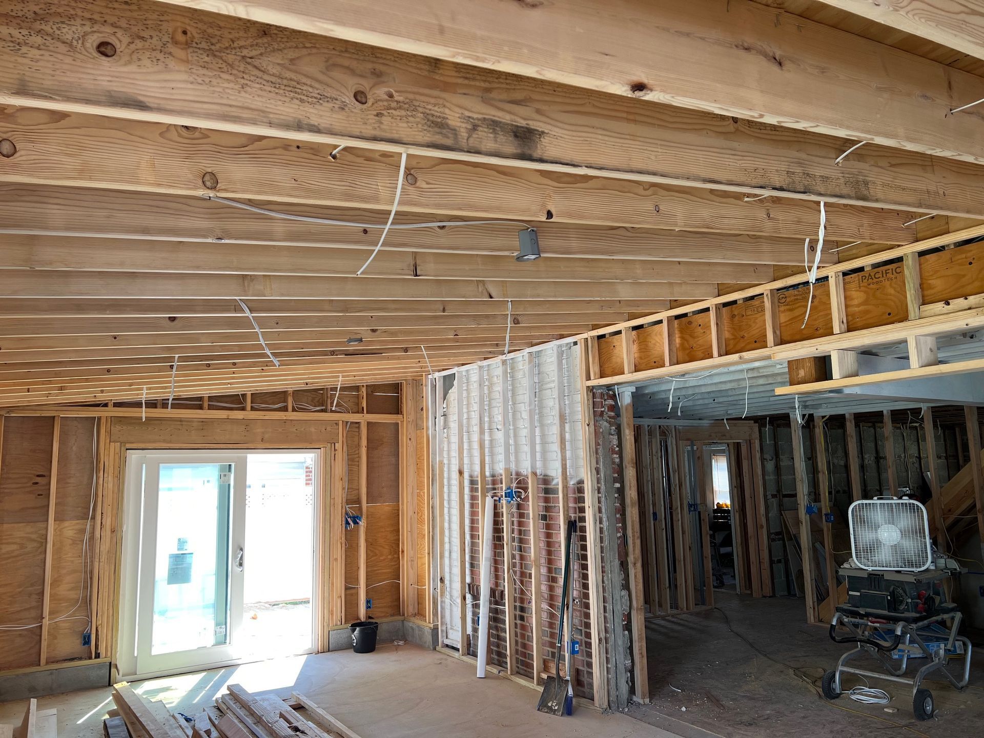 Interior view of unfinished house with exposed wooden beams, wiring, and framing. A sliding glass door is visible.