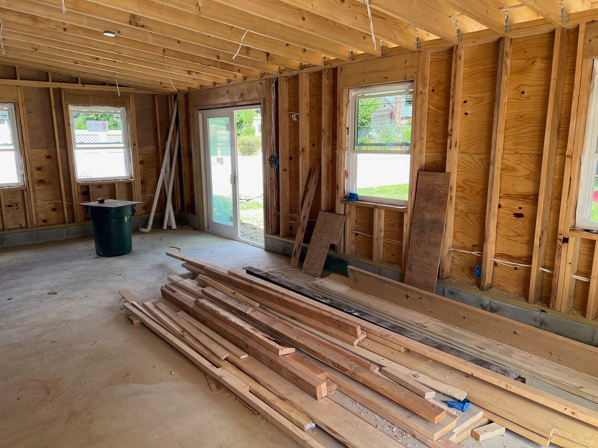 Interior of a room under construction, with exposed wooden framing, windows, and a sliding glass door.