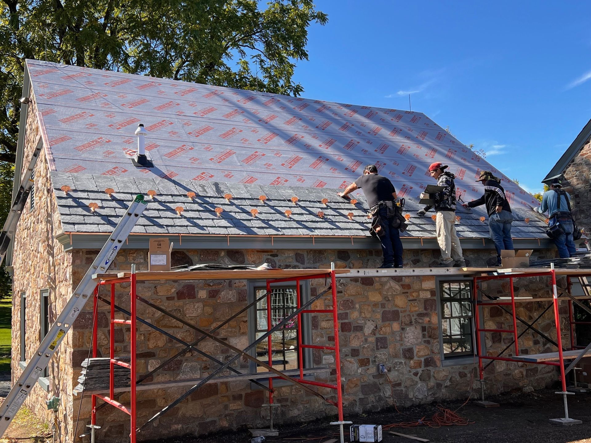 Roofers working on a stone building, installing a new roof on a sunny day. Scaffolding visible.