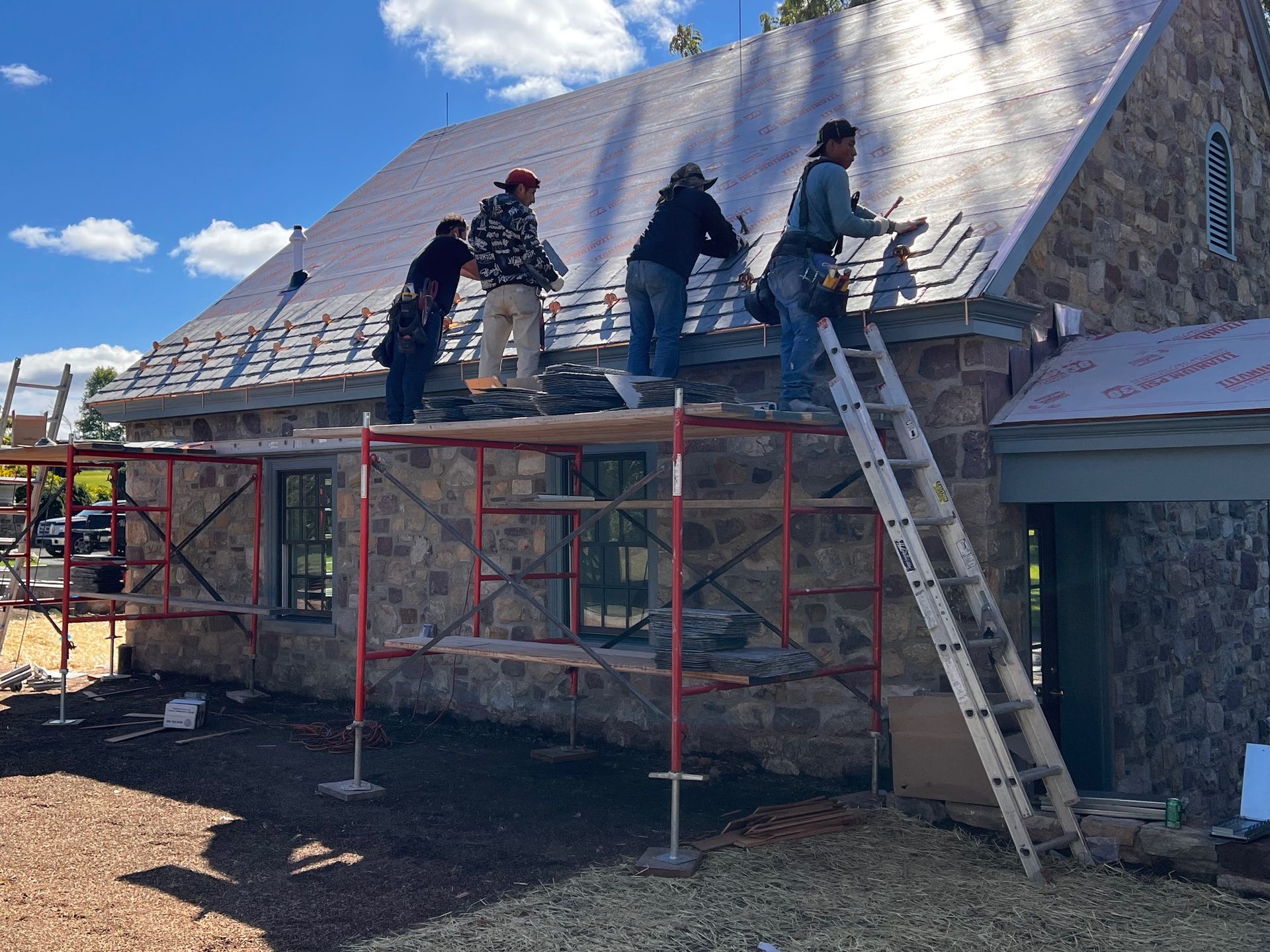 Construction workers on a stone building roof, using scaffolding and ladder on a sunny day.