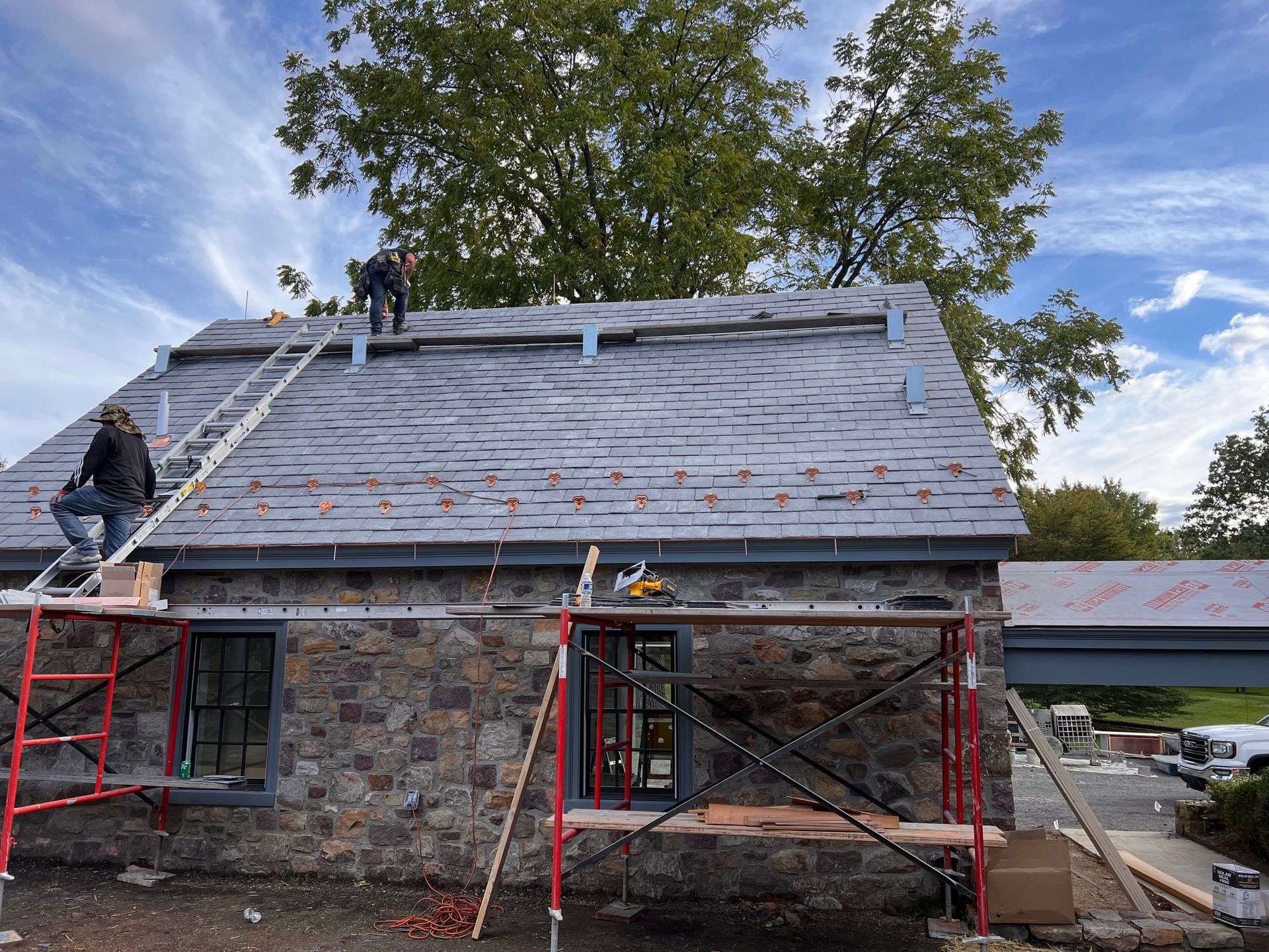 Roofers working on a stone building, installing new slate roof. Scaffolding, ladder, and tools visible. Blue sky.