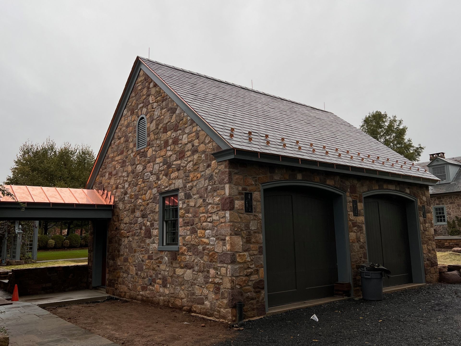 Stone garage with copper roof and side awning. Two dark garage doors, gray window frames, overcast sky.