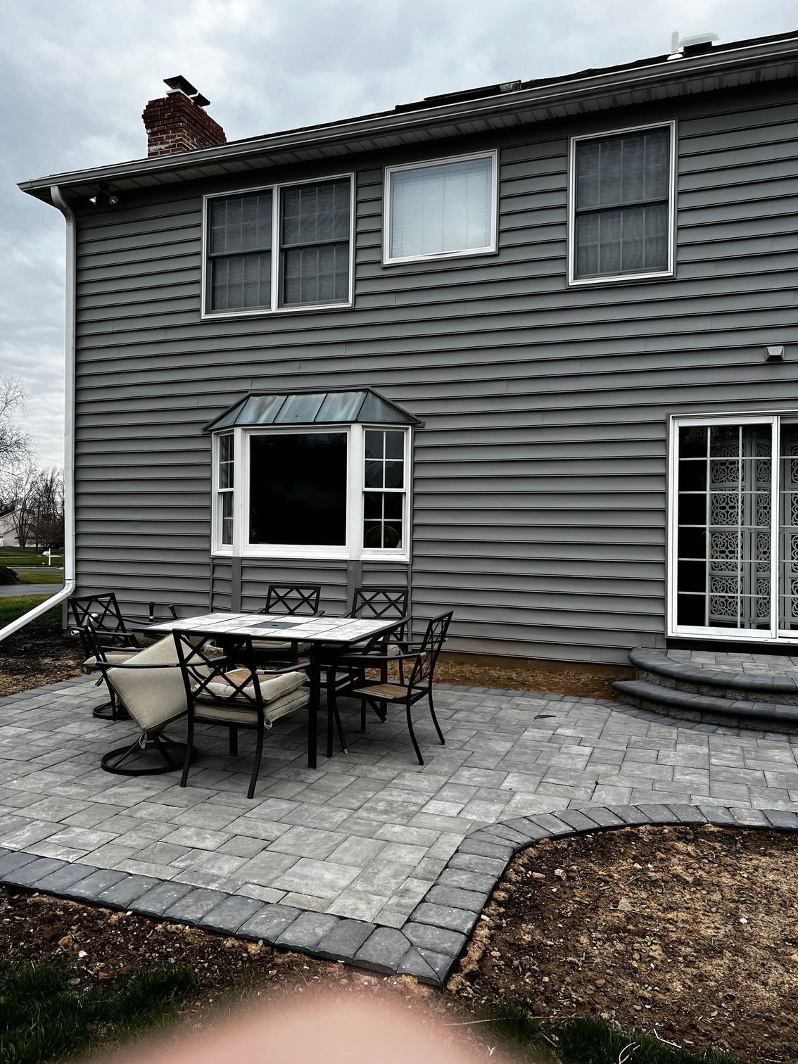 Back exterior of a gray-sided house with a patio and outdoor furniture; overcast sky.
