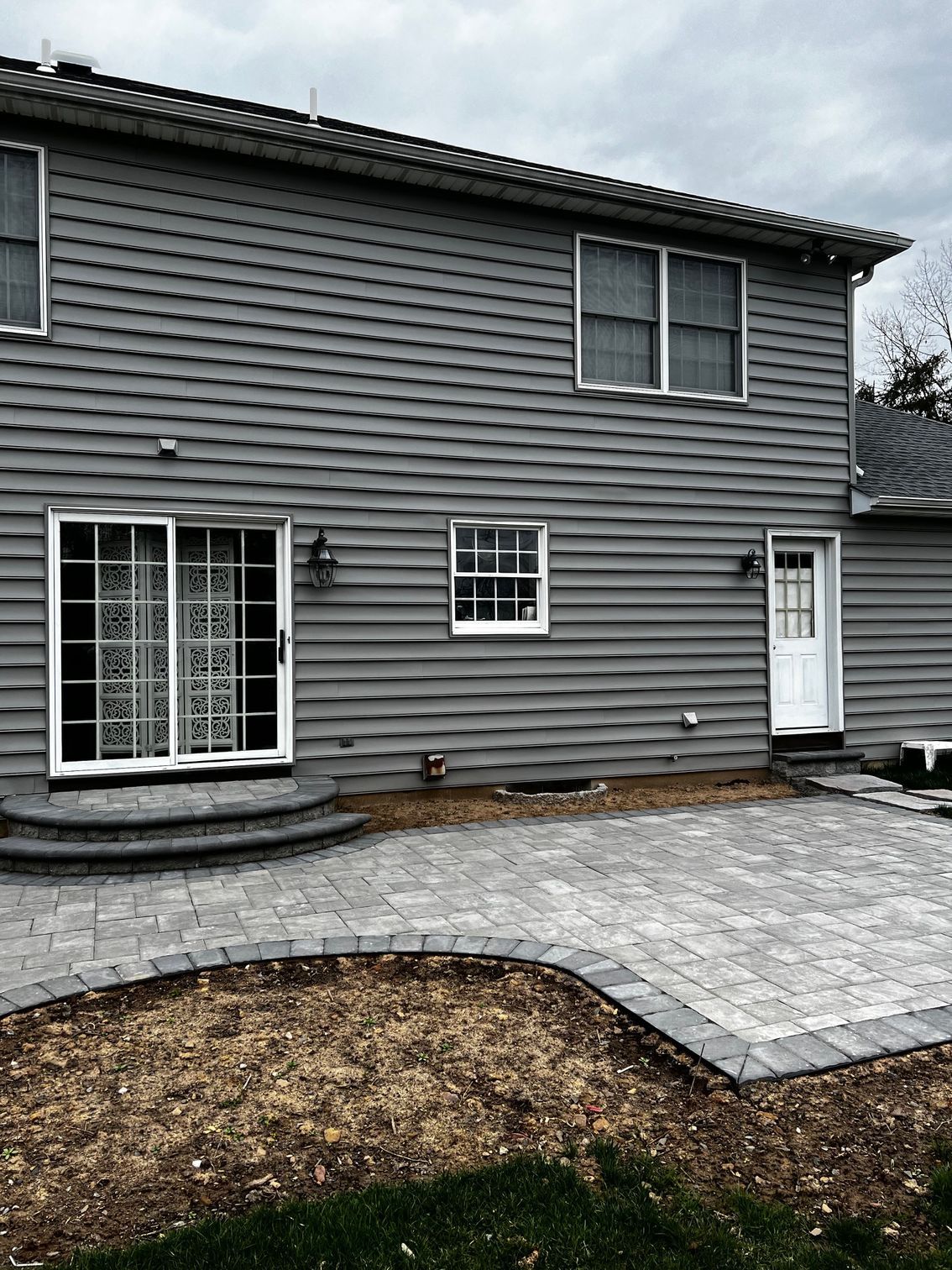 Gray house exterior with paver patio. Sliding glass door, small window, white door. Cloudy sky.