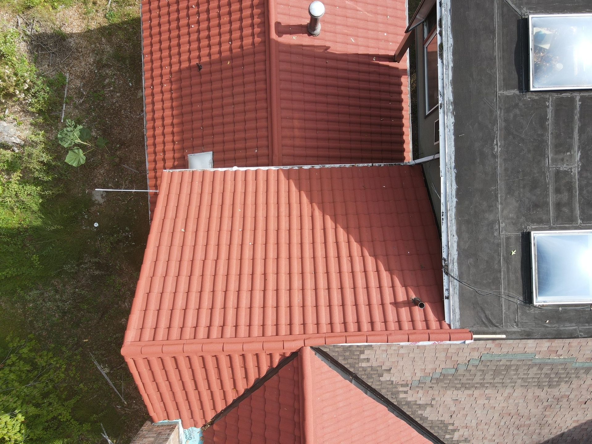 Overhead view of a house with red tile roofs and a flat black roof with skylights, next to green grass.