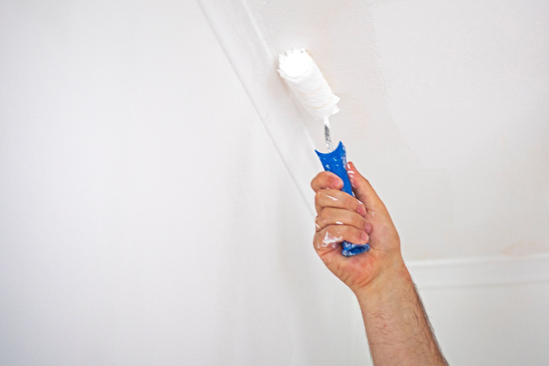 Person using a paint roller with blue handle to paint a white ceiling.