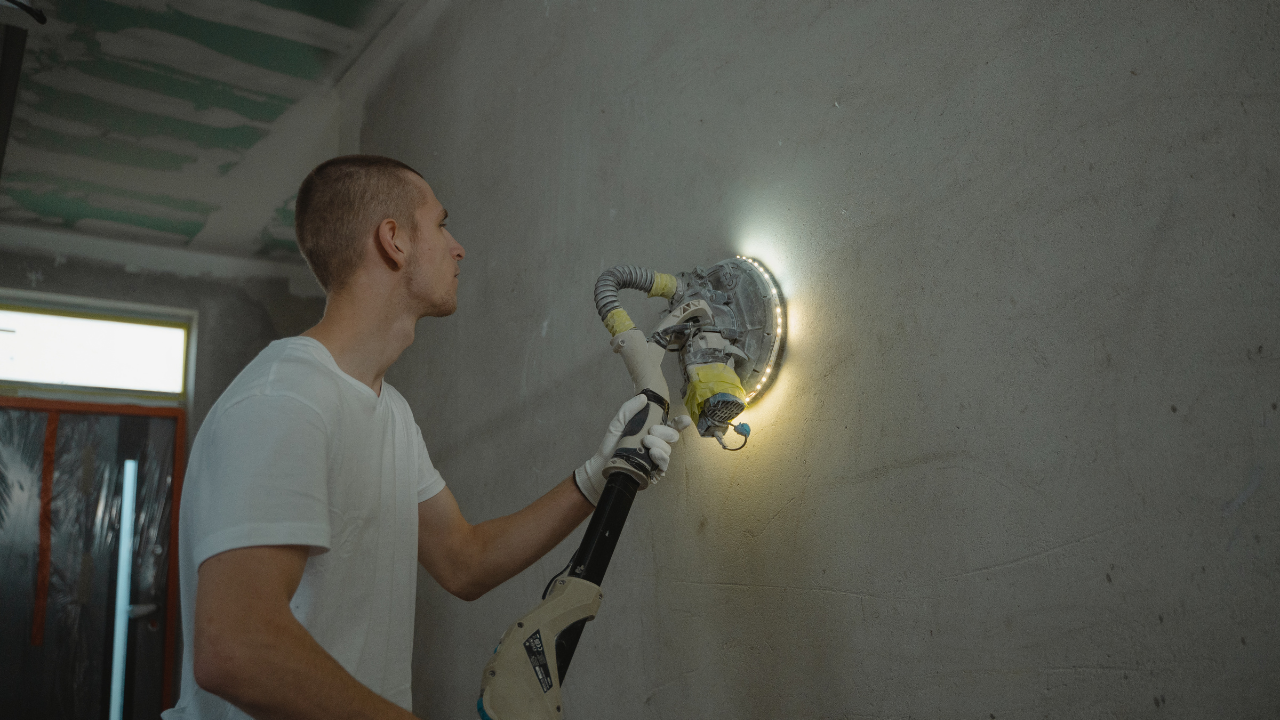 Man sanding a wall with a drywall sander, illuminated by a light.