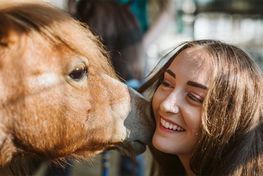 Woman smiling as a brown horse nuzzles her face outdoors