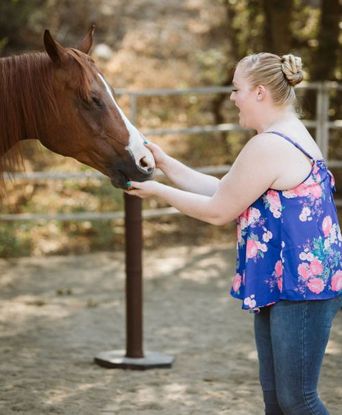 Woman in a blue floral top gently touching a horse’s face outdoors near a fence.