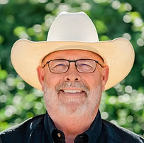Smiling person wearing a white cowboy hat and glasses against a blurred green background