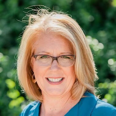 Smiling woman with blond hair and glasses outdoors against a green background