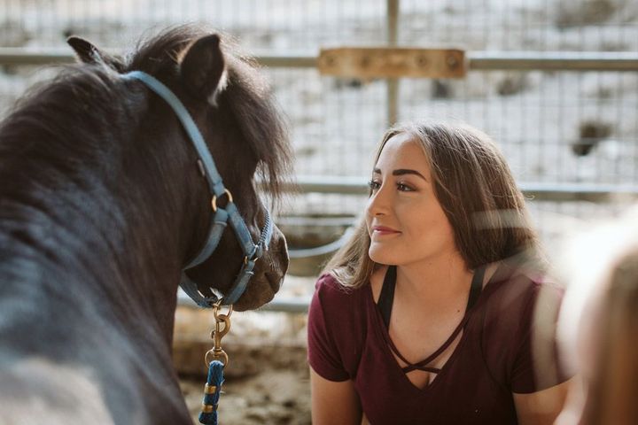 Woman kneeling beside a black horse in a fenced stable, looking at it with a gentle smile.