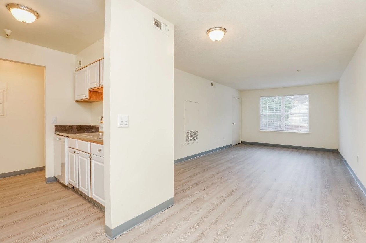 A bright apartment interior featuring a small kitchen with white cabinets next to an open living area with a large window.