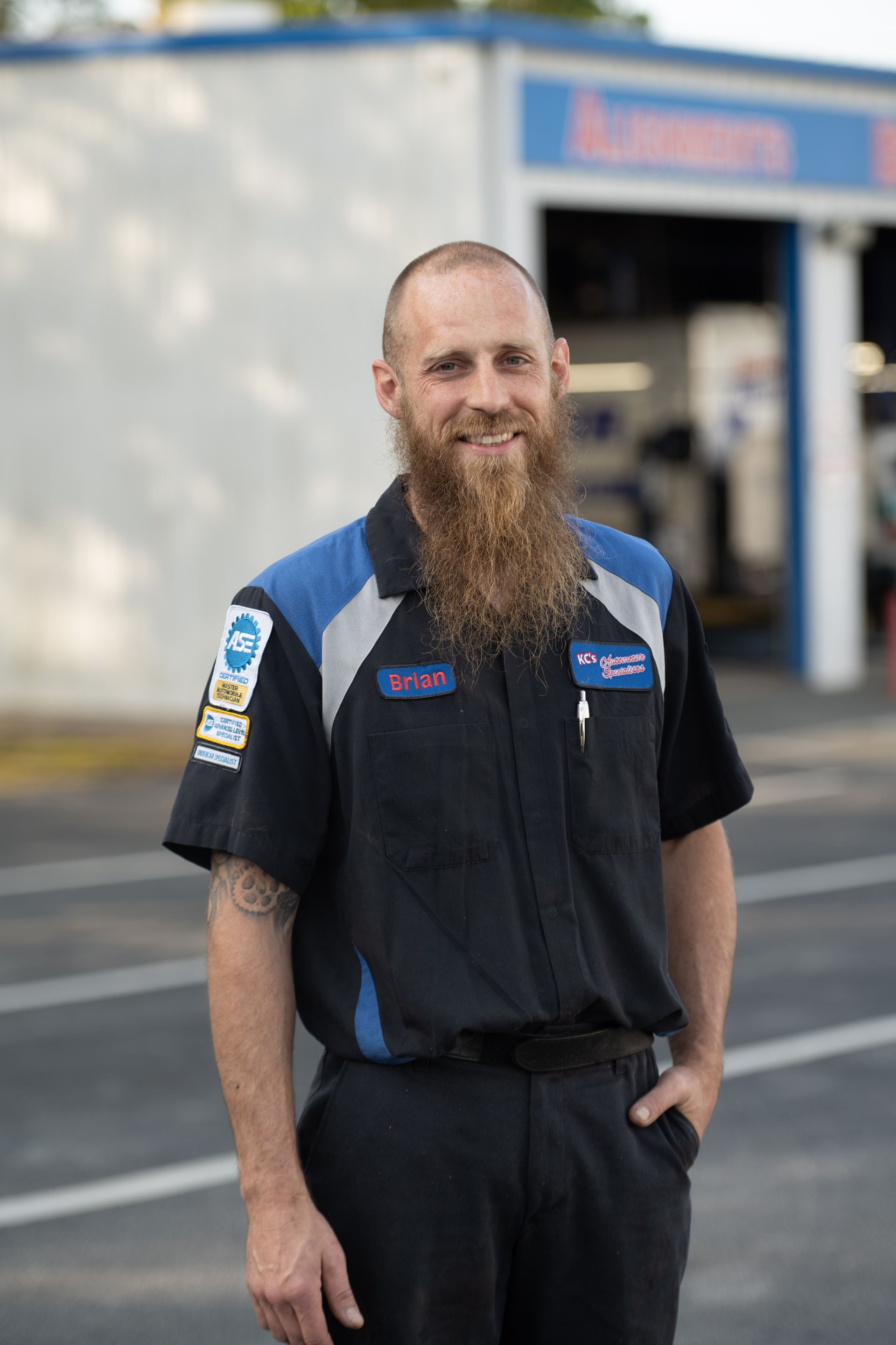 Mechanic with a long beard smiles in front of a garage. He wears a blue and black uniform with a patch.