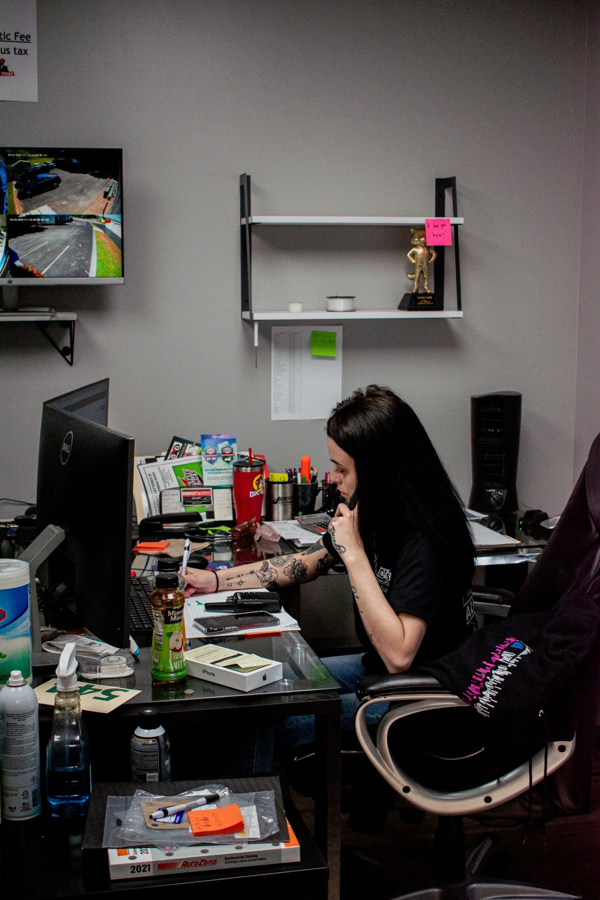 Person sits at desk, focused on work. Computer monitor, cluttered desk with food, drinks. Gray walls, shelves.