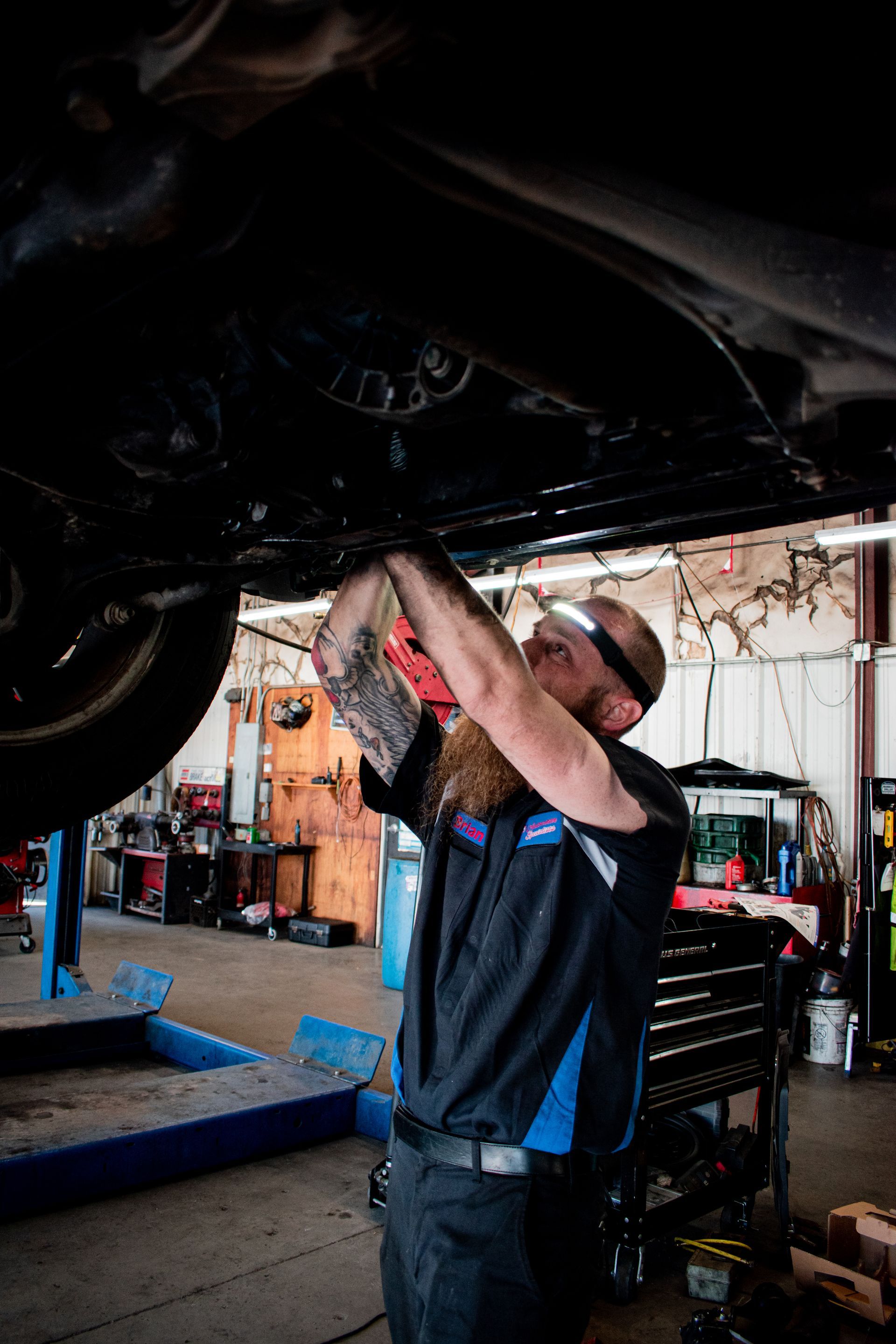 Mechanic working under a car, using a cloth. Shop interior with tools, overhead light, and a lift.