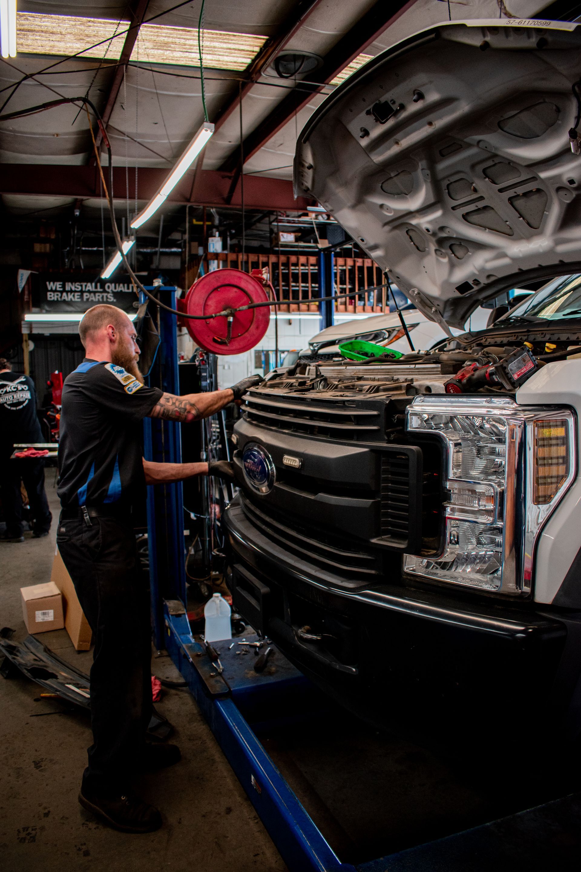 Mechanic working on the front of a truck inside an auto repair shop. Hood is open, tools visible, and a lift is supporting the truck.