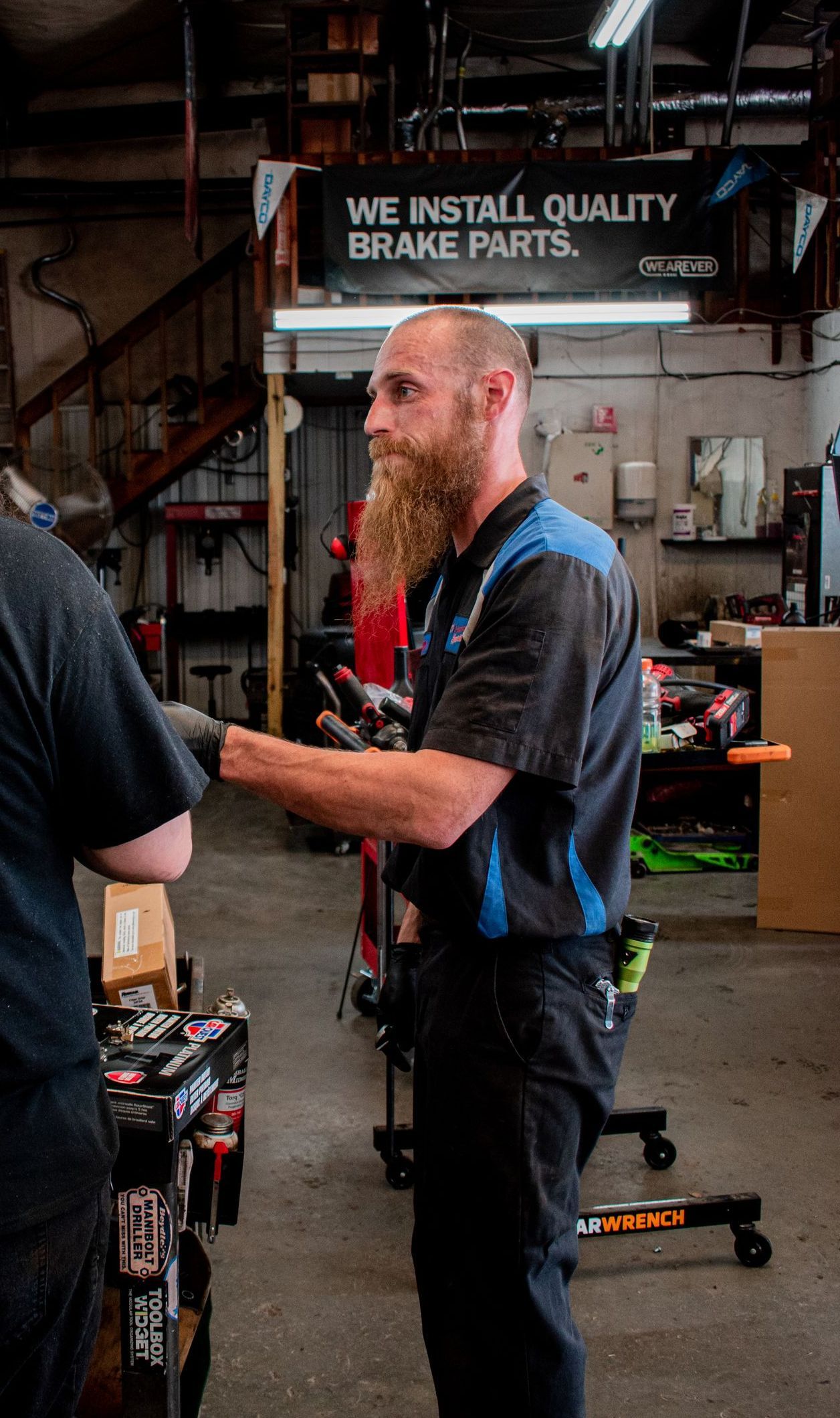 Mechanic with long beard in auto repair shop, holding tools, working on vehicle. Shop interior, sign: 