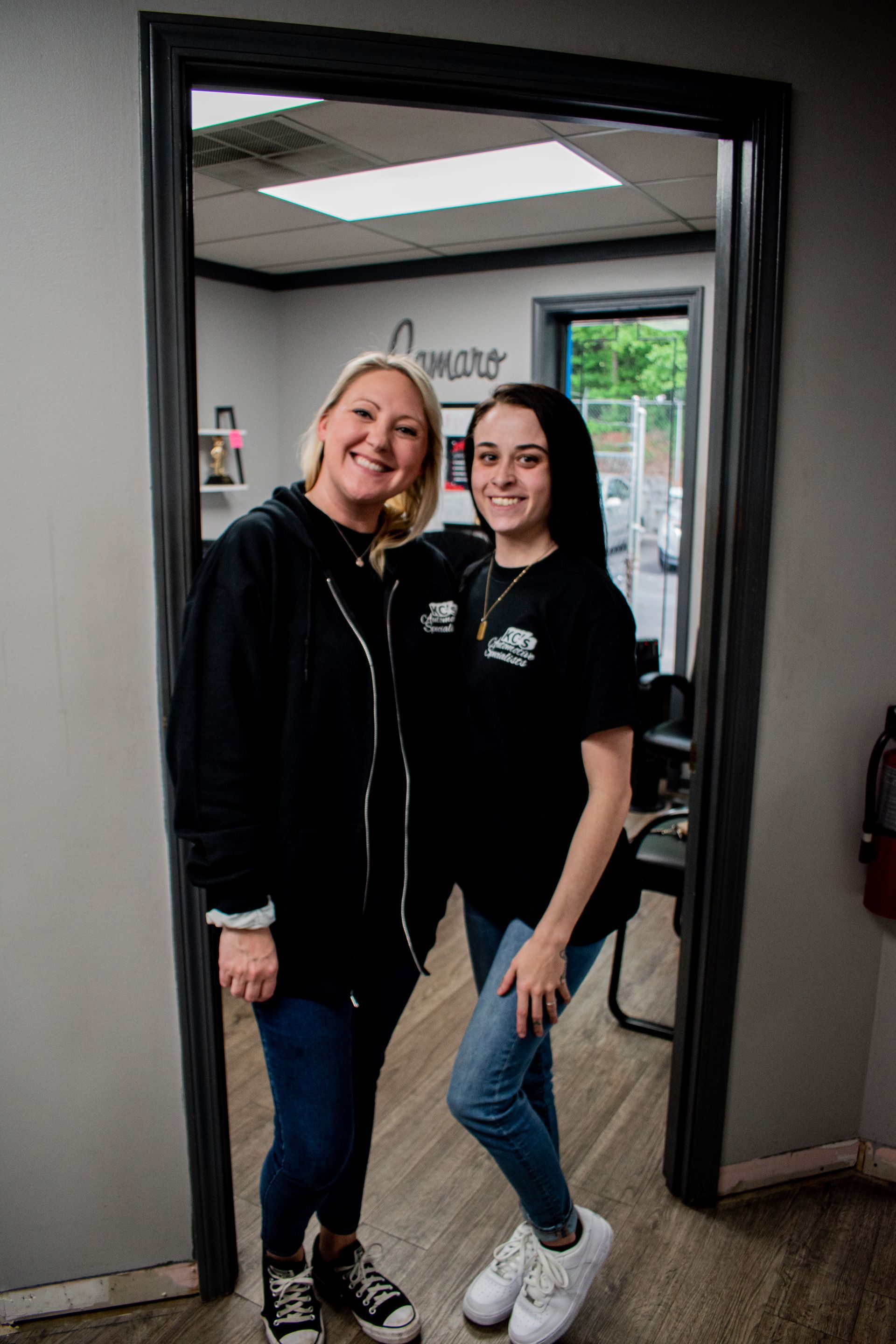 Two women inside a doorway: one blonde, one with dark hair. Both smile, pose, wear casual clothes.