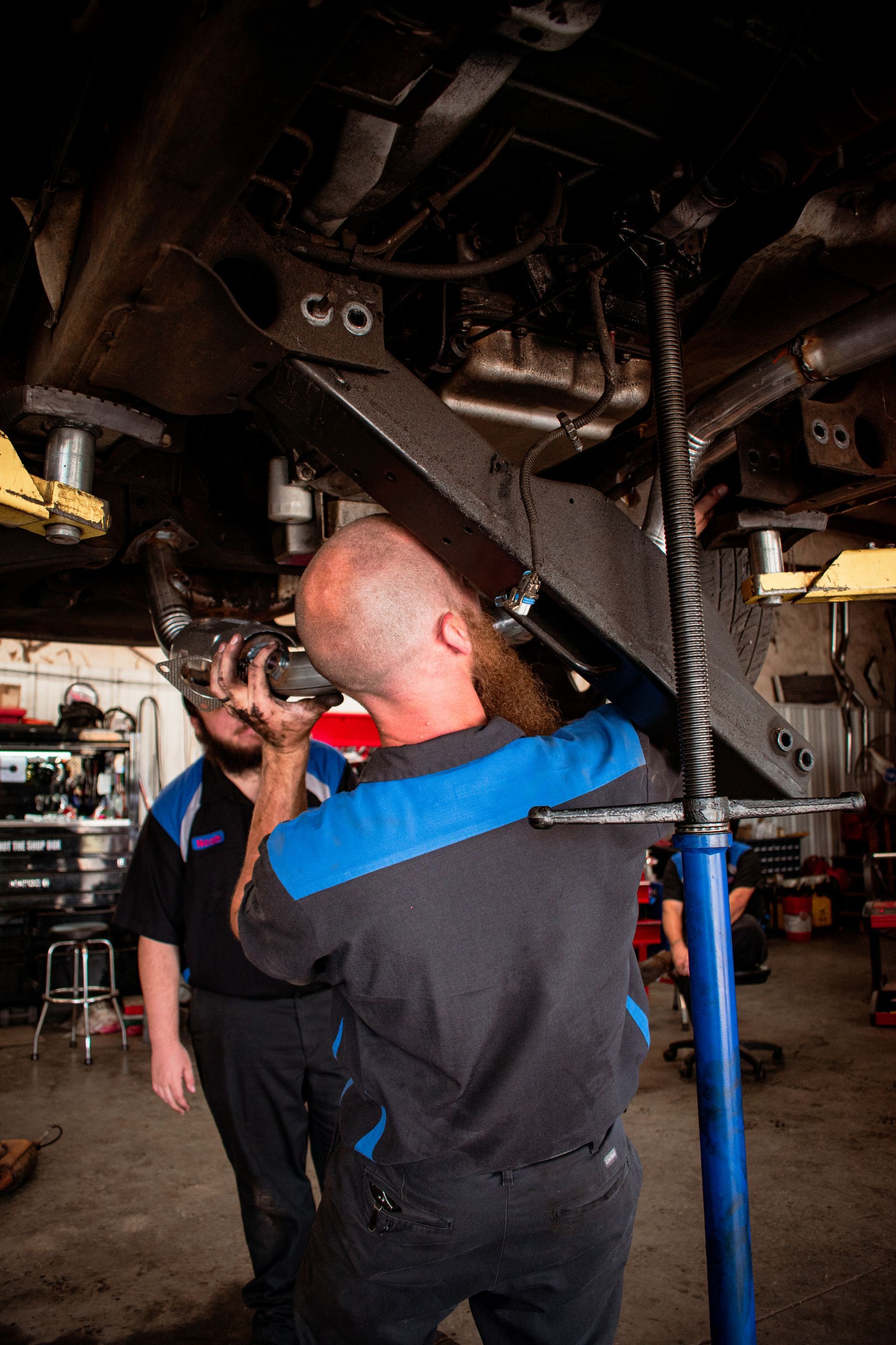 Mechanics working on the undercarriage of a vehicle in a garage. One uses tools, another observes.