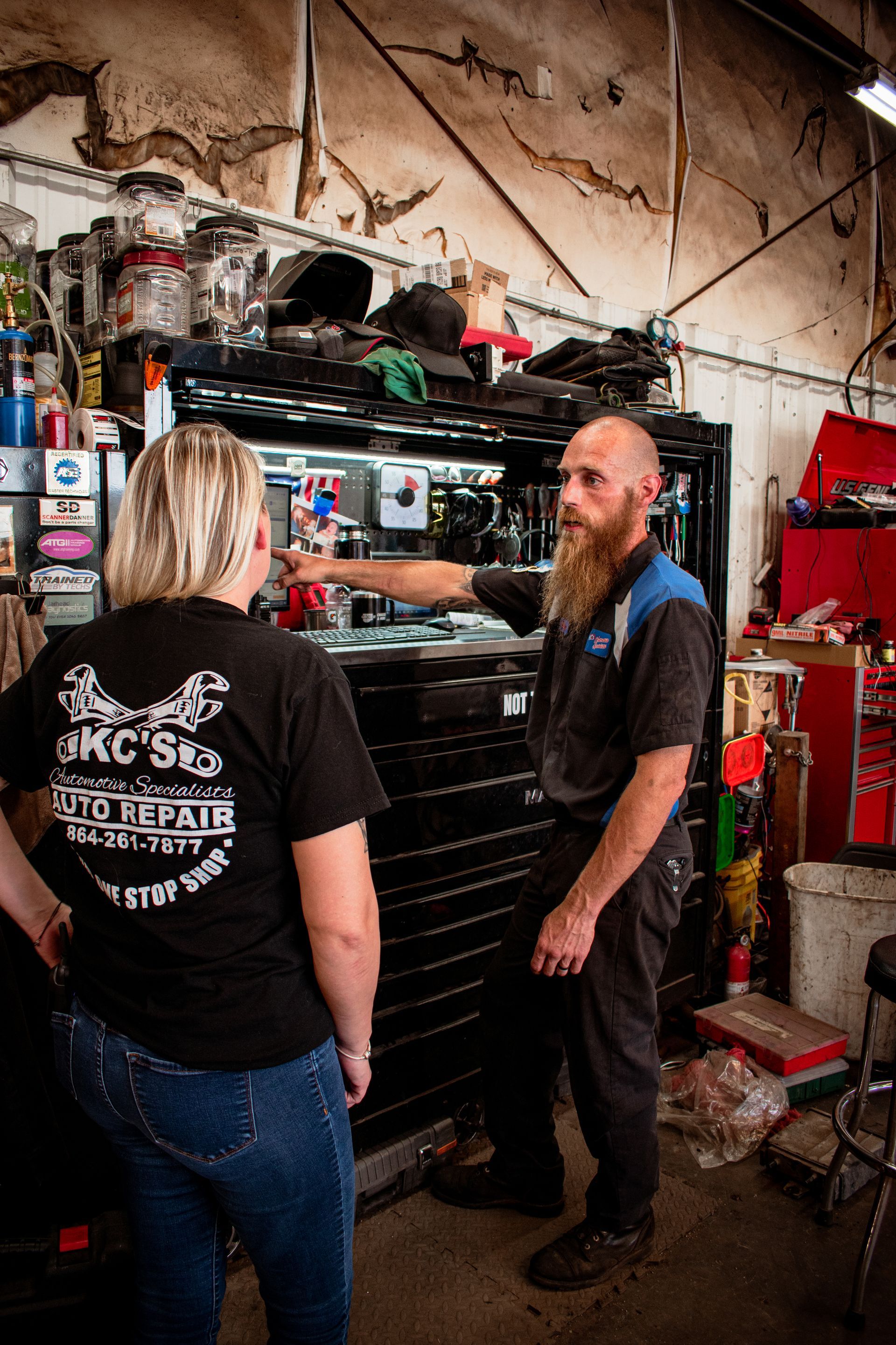 Mechanic in blue and black work clothes gestures towards black toolbox, explaining something to a person in black KC's shirt and jeans in a garage.