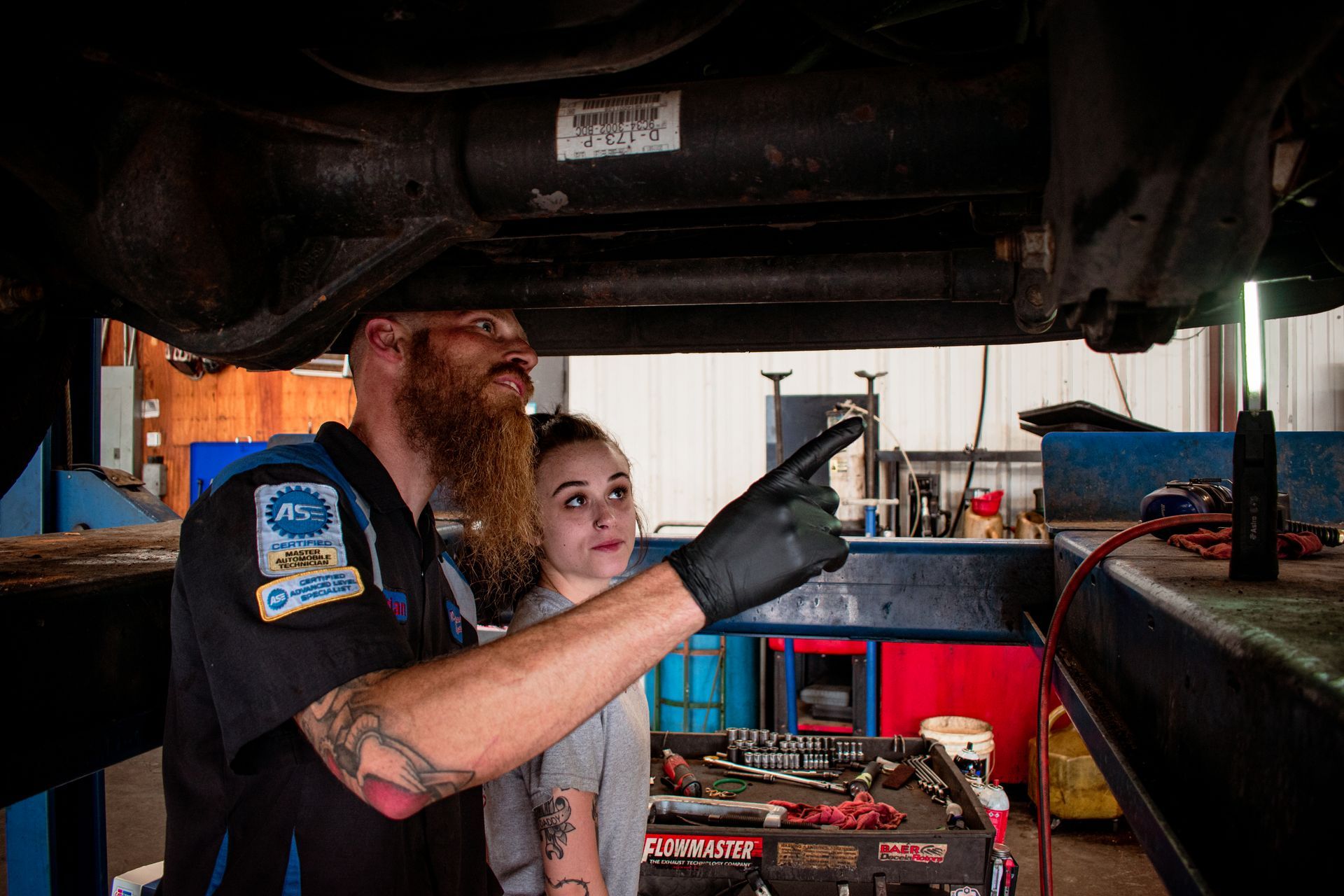 Mechanic points to car part while explaining to a customer under a vehicle. Workshop setting.