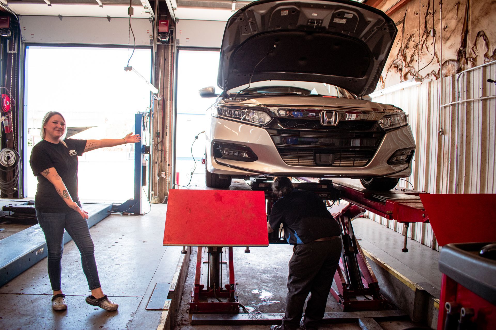 A person points at a silver car on a lift in a garage. Mechanic works underneath.
