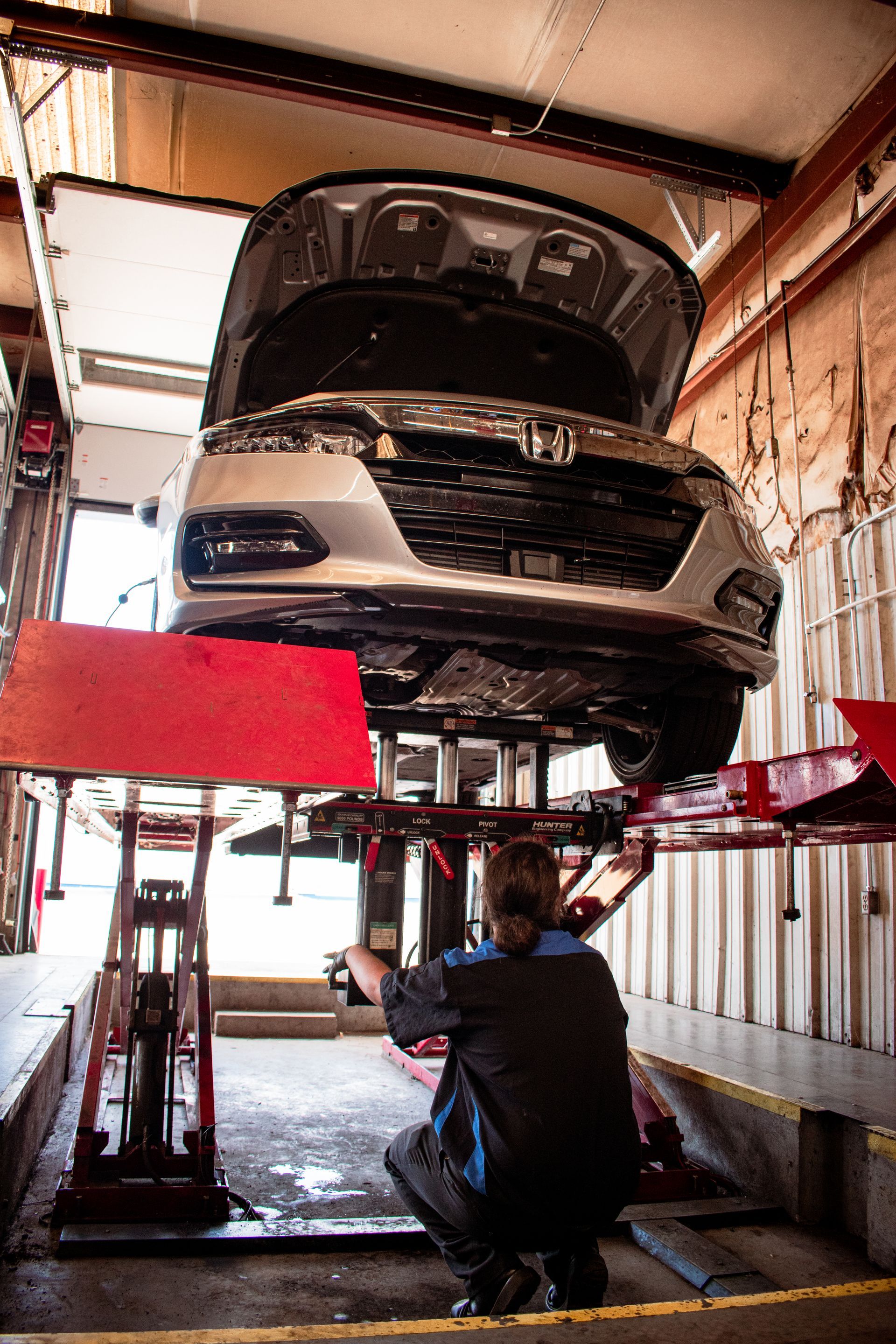 Mechanic crouches under a car on a lift, inspecting the underside of the vehicle in a garage.