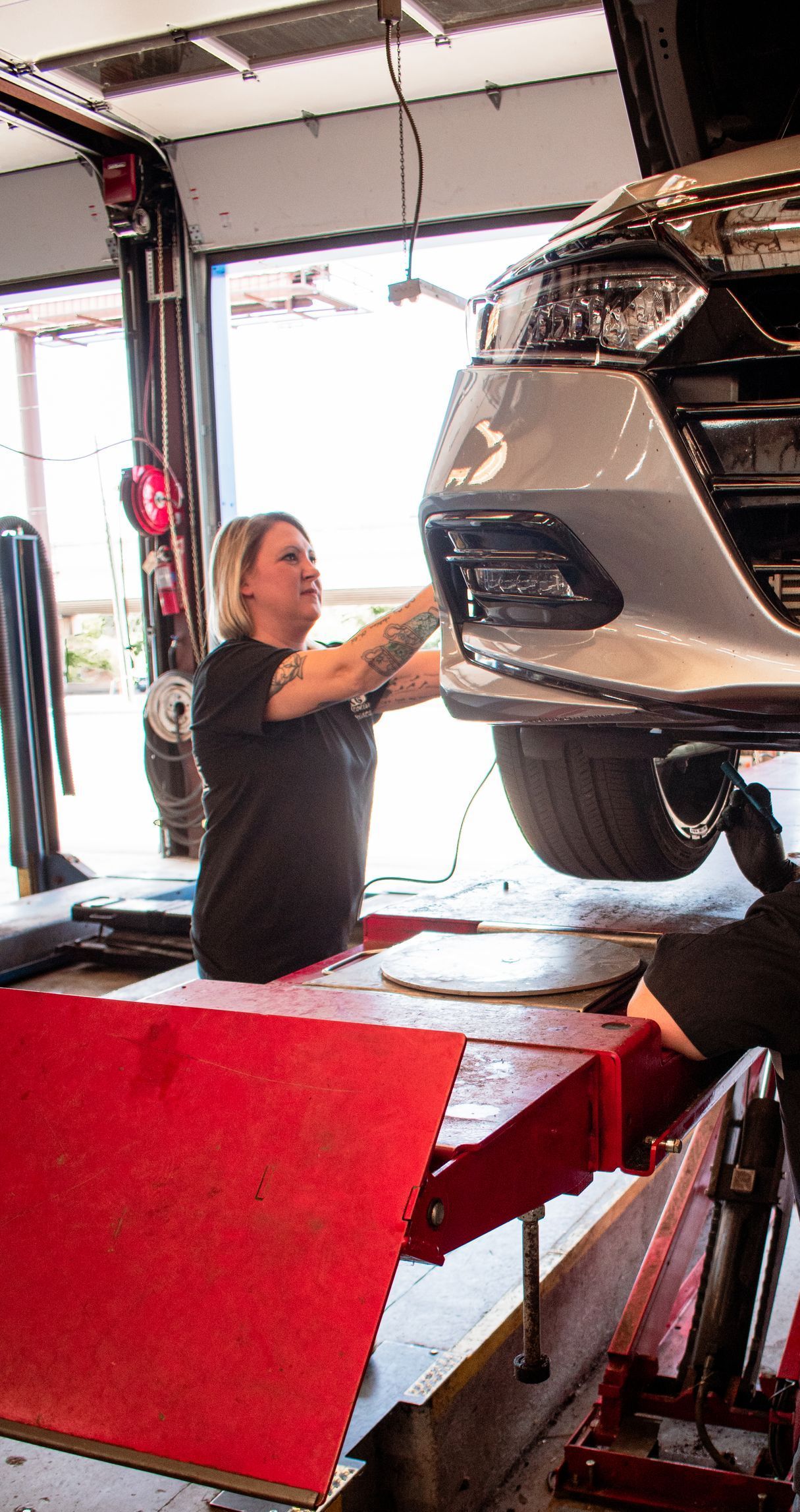 Woman in a black shirt working on a car raised on a lift in a garage.