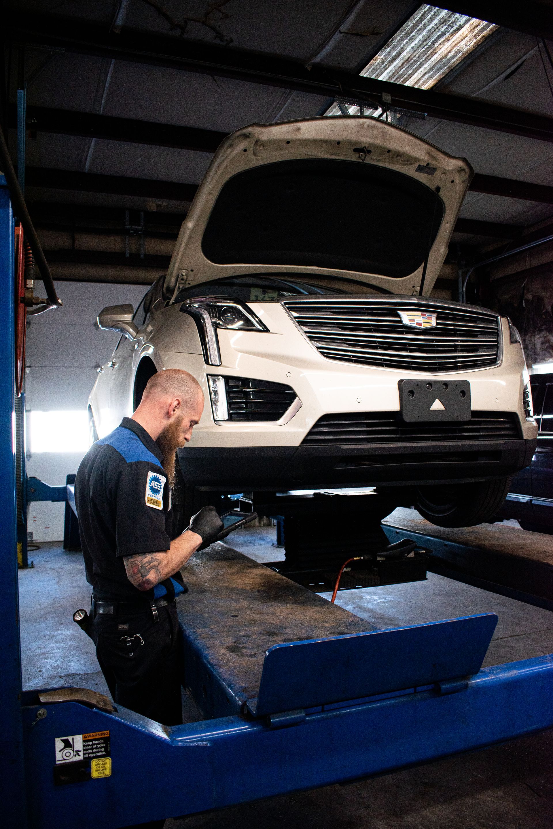 Mechanic examining underside of a white car lifted in a shop. Mechanic wearing a black and blue uniform. Shop setting.