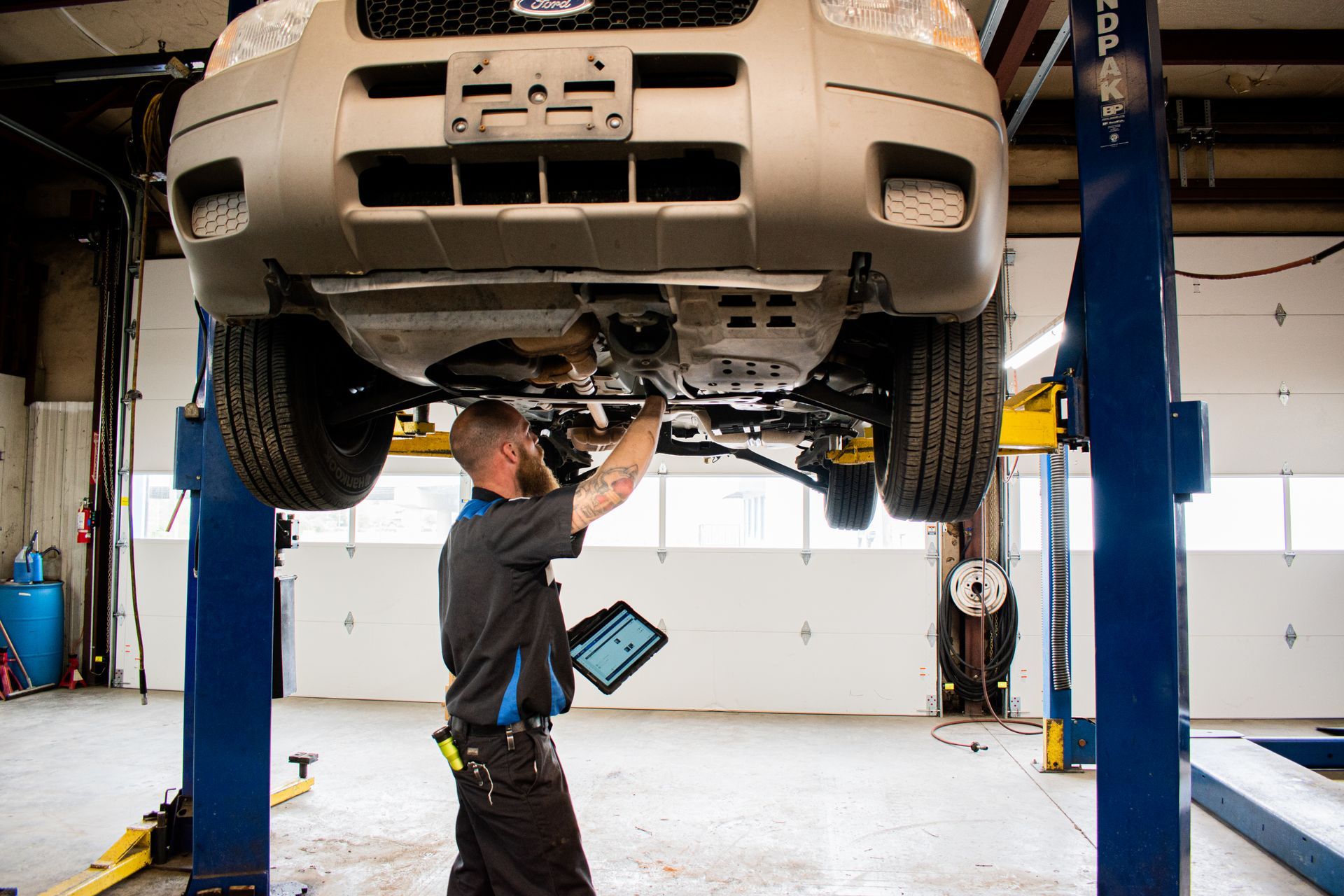 Mechanic working on the underside of a car raised on a lift in a garage, using a tablet.