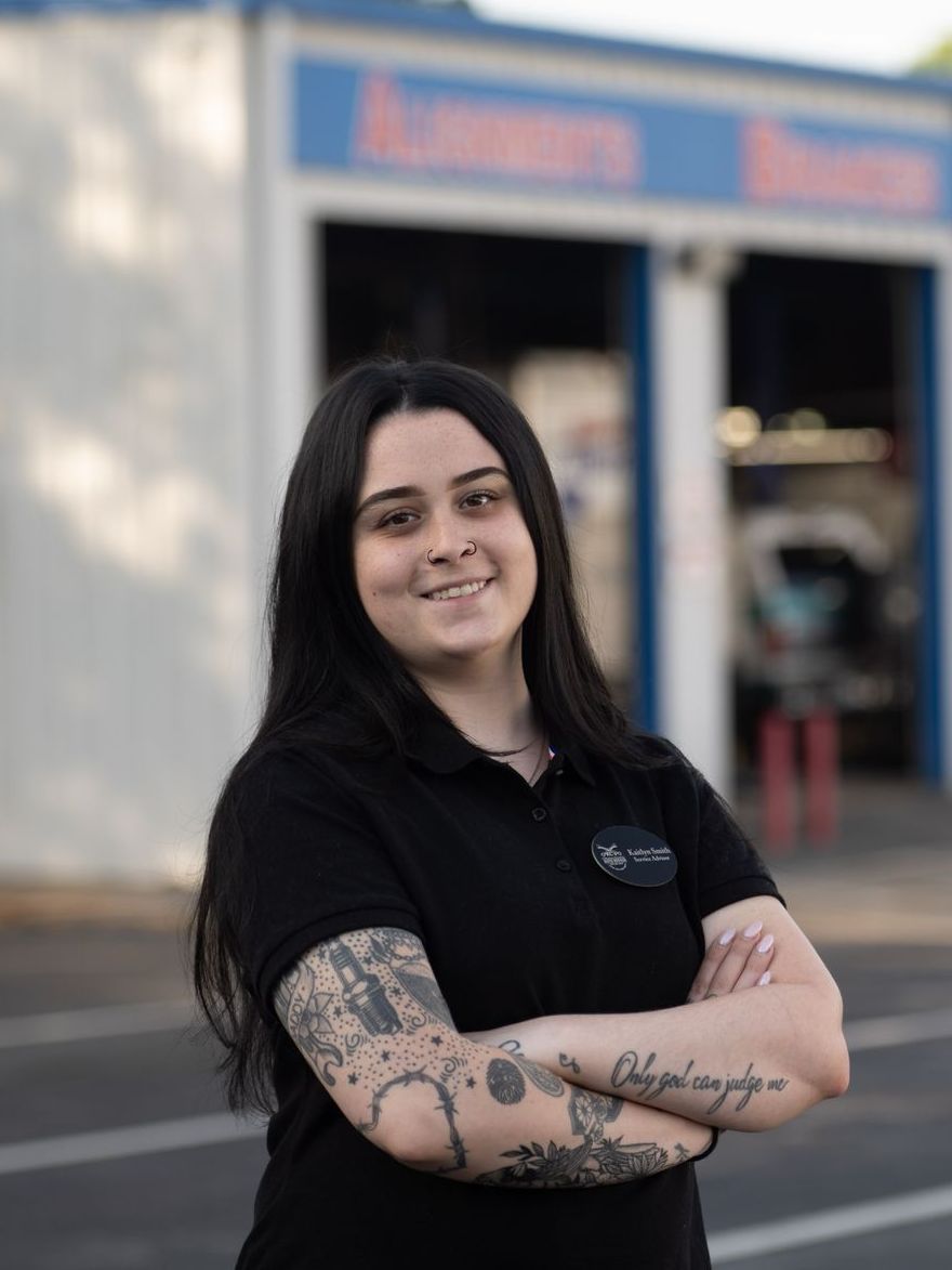 Woman with tattoos stands in front of a garage, arms crossed, smiling.
