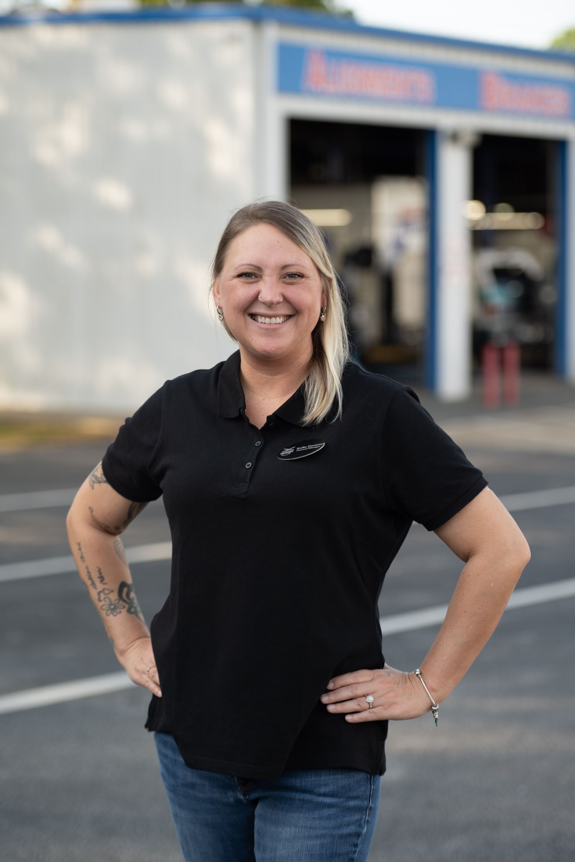 Woman in black shirt and jeans smiling in front of auto repair shop.