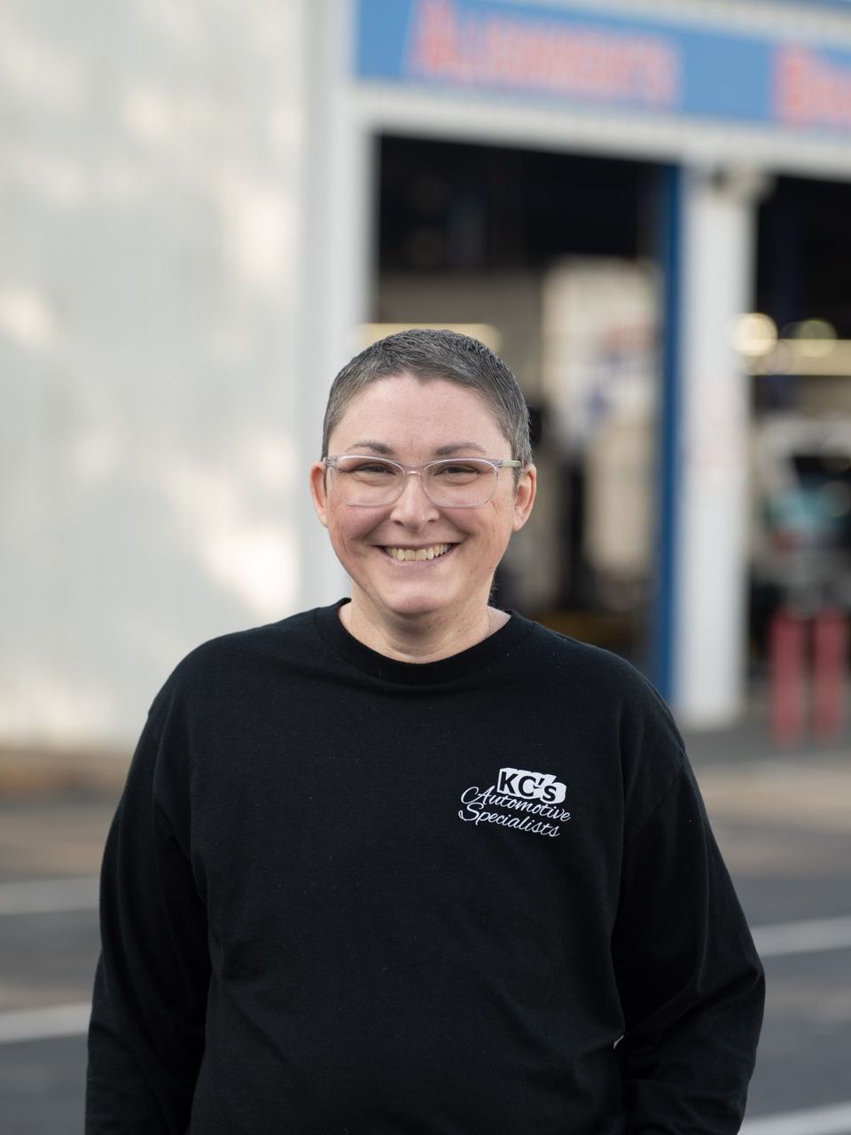 Woman with short hair smiles, wearing glasses and a black sweatshirt, standing outside an auto repair shop.