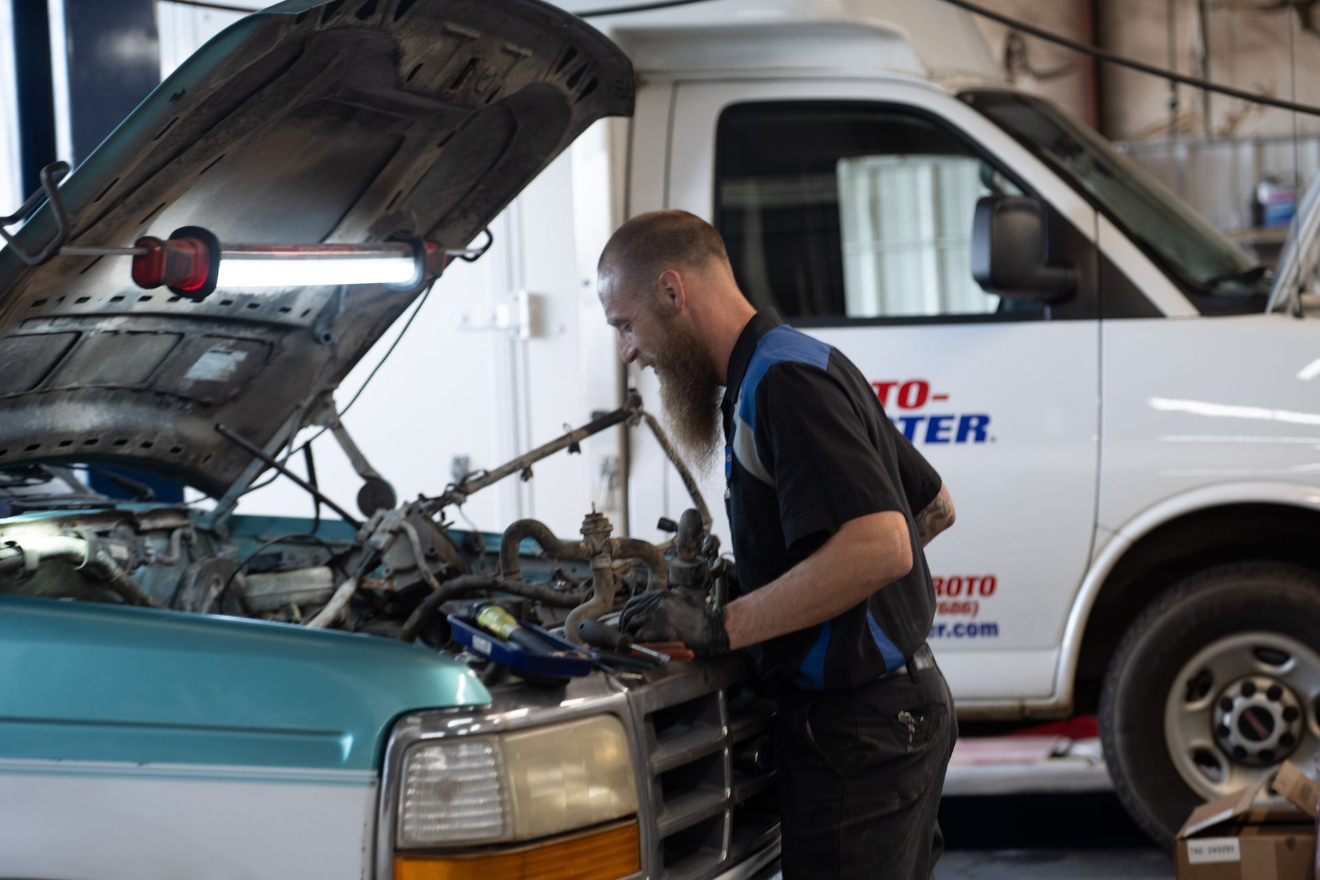 Mechanic with beard working on a car engine in an auto shop with a white van.