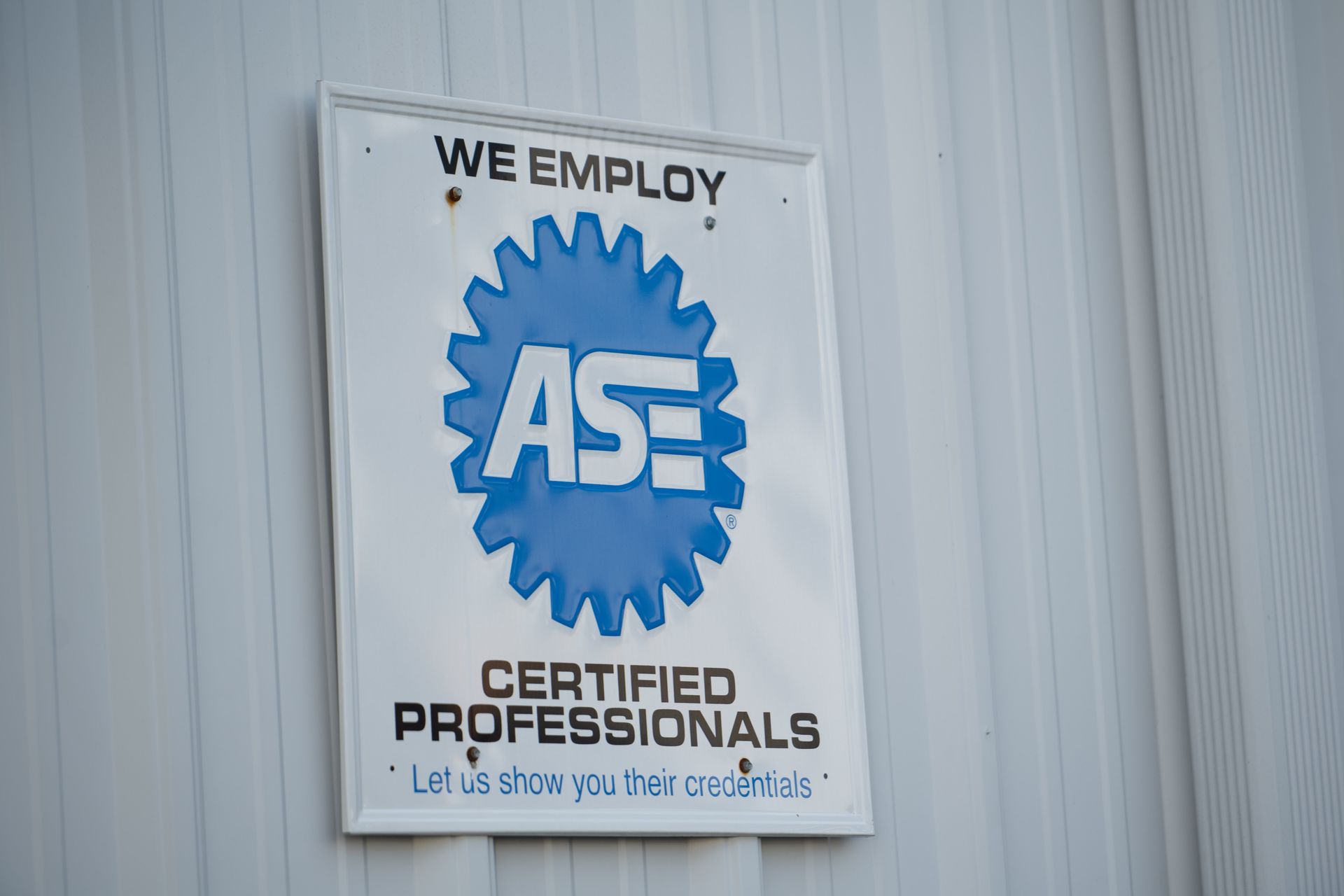 Sign indicating a business employs ASE certified automotive professionals. White sign, blue gear with ASE logo.