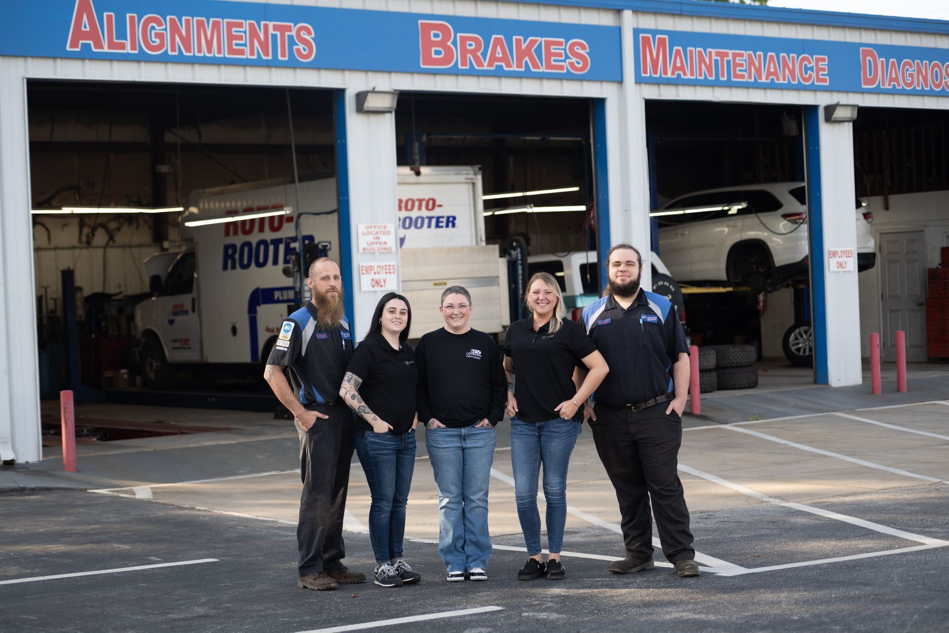 Five people standing in front of an auto repair shop. Blue and white building with car up on a lift.