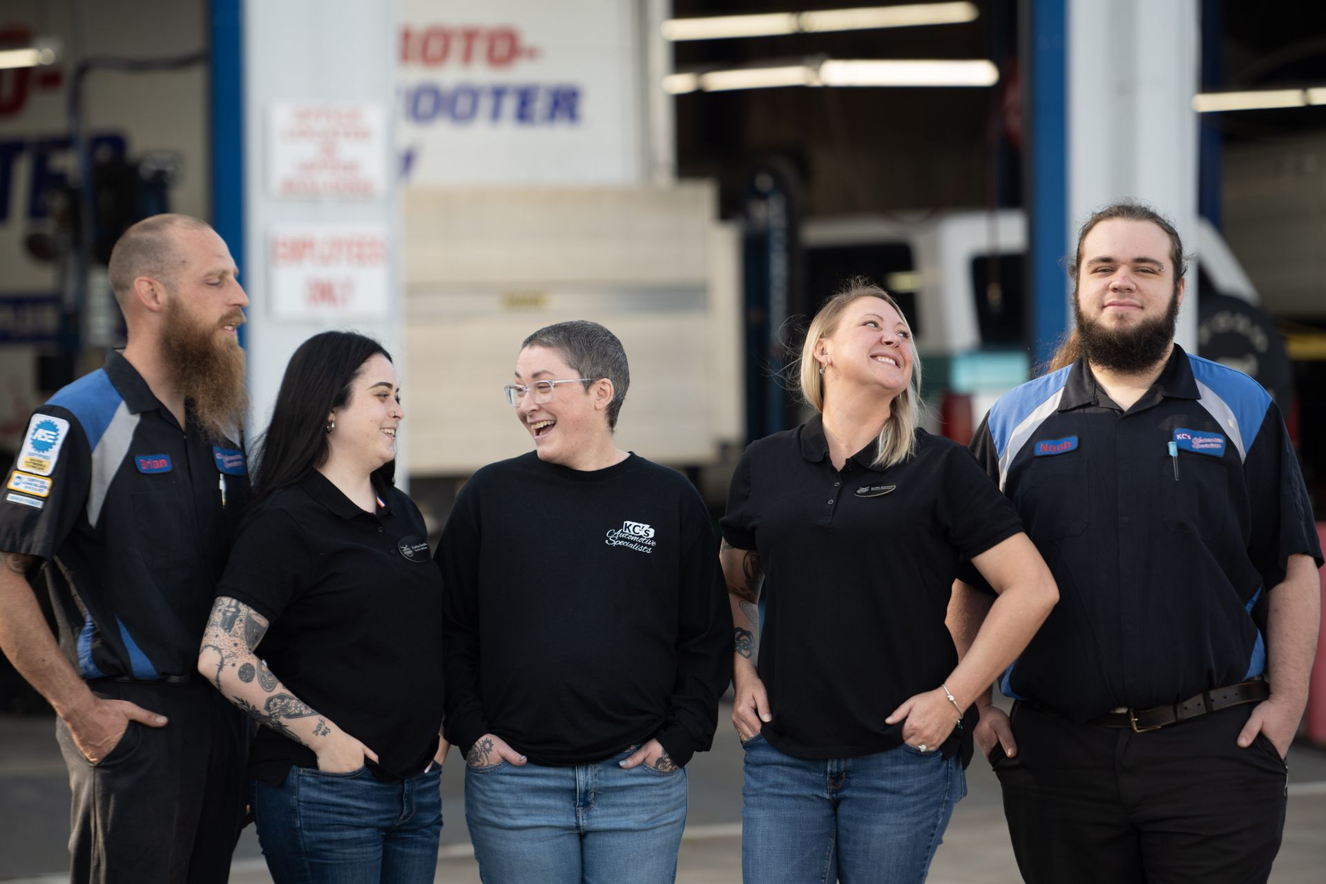 Team of five mechanics at an auto shop, smiling. Two men, three women, wearing work shirts. Blue and black colors.