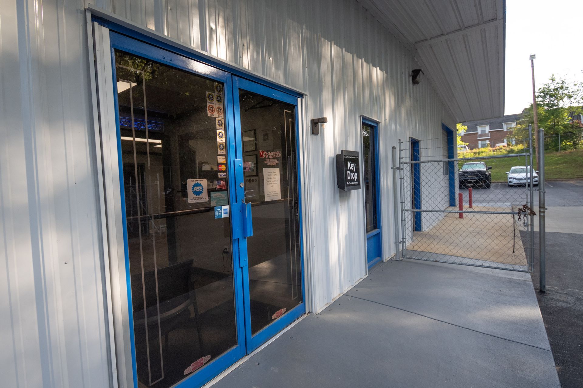 Exterior of a building with blue-framed glass doors and a sidewalk entrance. Gray and white siding. A mailbox is visible.