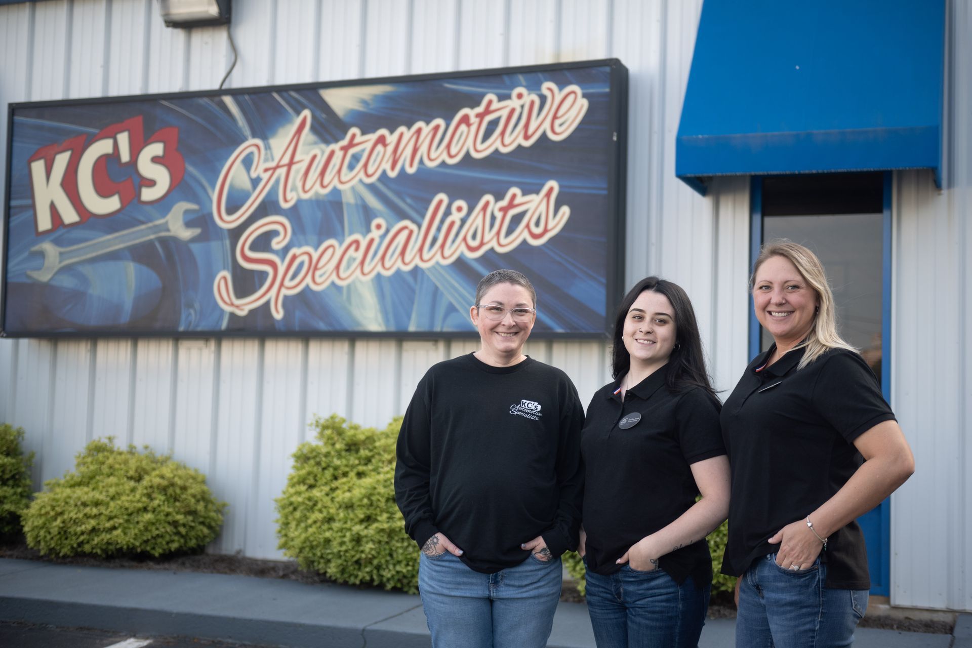 Three women stand in front of KC's Automotive Specialists sign. The women are smiling and wearing black shirts.
