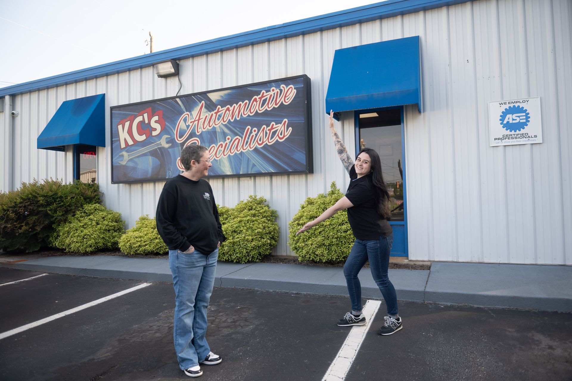 Two people outside KC's Automotive Services. One points, the other looks at them. Blue awnings, a sign, and shrubs are present.